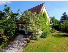 Ein Einfamilienhaus mit rotem Ziegeldach und teilweise mit Efeu bewachsener Fassade, umgeben von einem gepflegten Garten mit Rasen, Bäumen und Büschen. Ein gepflasterter Weg führt zur Eingangstür, und im Hintergrund sind weitere Grünflächen und ein Gartenstuhl erkennbar.