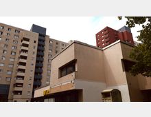 Wohnkomplex mit mehreren mehrstöckigen Gebäuden in schlichten Farben, darunter beiger und roter Putz sowie Balkone. Im Vordergrund ein flaches, hellbeiges Gebäude mit großen Fenstern und einem Baum an der rechten Bildseite.