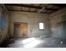 Leerer, unfertiger Innenraum eines Lager- oder Werkstattraums mit rohen, verputzten Wänden, Betonboden und sichtbar konstruiertem Dach mit Holzträgern und einer hängenden Leuchtstofflampe. An der Rückwand befinden sich eine provisorische Tür mit Holzplatten und ein kleines Fenster; am Boden liegen einige Säcke und Baustellenmaterial.