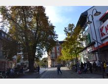 Straßenszene in einer Fußgängerzone mit Kopfsteinpflaster, umgeben von Bäumen und Gebäuden. Links sind kleinere Geschäfte und Sitzbänke sichtbar, rechts ein modernes Geschäft mit Schildern, sowie Menschen, Fahrräder und Kinderwagen.