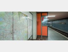 U-Bahn-Station "Parchimer Allee" mit blau gefliester Wand, orangefarbenen Verkleidungen, einer markanten Rohrsäule und einer aufgestellten Karte. Der Bahnsteig ist unbesetzt, und das Gleis erstreckt sich nach rechts.