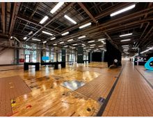 Weitläufige Innenansicht einer Industriehalle mit hellem Holz- und Fliesenboden. Die Decke zeigt freiliegende Metallträger und Beleuchtung, während schwarze Trennwände und minimalistische Tische im Raum verteilt sind.