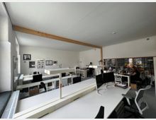 Großraumbüro mit weißen Arbeitsplätzen, mehreren Monitoren und Regalen. Der Raum hat eine helle Gestaltung mit Holzbalken an der Decke, großen Fenstern und einigen Wanddekorationen, wie gerahmte Bilder.