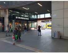 Eine überdachte Passage an einem Bahnhof mit einem Starbucks-Café auf der linken Seite. Im Hintergrund ist eine gelbe Straßenbahn sowie ein Bereich mit Glasfassaden und Treppen zu sehen, während Passanten durch die Szene gehen.