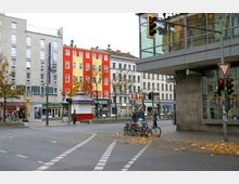Straßenecke in einer Stadt mit mehrgeschossigen Wohnhäusern und Geschäften im Erdgeschoss. Im Vordergrund eine Kreuzung mit Fahrradständern, Fußgängern und Ampeln; im Hintergrund bunte Fassaden und ein Eingang zur U-Bahn.