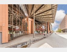 Außenansicht der Spielbank Berlin am Potsdamer Platz mit moderner Glasfassade, überdachtem Eingangsbereich und säulenförmigen Stützen; im Vordergrund ein weitläufiger, gepflasterter Platz mit Geländern entlang eines Wasserlaufs.