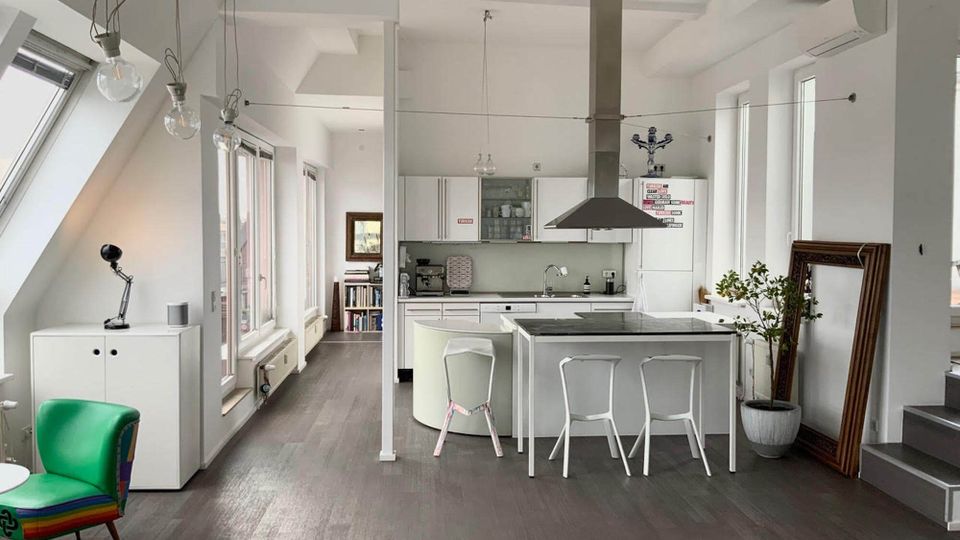 Bright open-plan loft kitchen and dining area with white cabinetry, a central island with stools, and a large metal range hood. Tall windows and slanted skylights line the left wall, with exposed hanging bulb lights, dark wood flooring, and a staircase on the right.