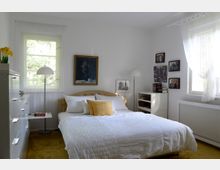 Helles Schlafzimmer mit einem Doppelbett, weißer Bettwäsche und gelbem Zierkissen, umgeben von weißen Wänden. Links ein hohes Fenster mit Vorhang, daneben eine Kommode und eine Stehlampe; an den Wänden hängen Bilder und über dem Bett ist ein Gemälde angebracht.
