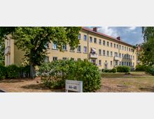 Mehrstöckiges Wohngebäude mit gelber Fassade, weißen Fenstern und kleinen Balkonen, umgeben von Bäumen, Sträuchern und einer Wiese. Im Vordergrund steht ein Schild mit der Aufschrift „Maxim-Gorki-Strasse 44-46“.