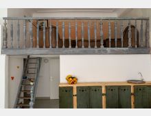 Raum mit einer offenen Holzempore, einer rustikalen Holztreppe und einer Wand mit weißem Anstrich. Unten befindet sich ein hölzernes Sideboard mit grünen Türen und darauf eine Schale mit Obst.