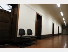 Ein schlichter Korridor mit cremefarbenen Wänden, Holzrahmentüren und einer glänzenden braunen Bodenfläche; zwei gepolsterte Stühle stehen an der Wand, Deckenleuchten sorgen für gleichmäßige Beleuchtung.