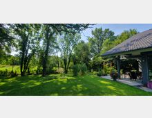 Garten mit einer Rasenfläche und hohen Bäumen im Hintergrund, die Schatten spenden. Rechts im Bild ist eine Terrasse mit Überdachung, Gartenmöbeln und blühenden Pflanzen in Töpfen zusehen.