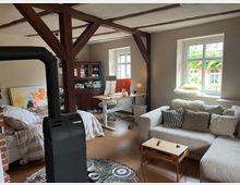 Offener Wohnraum mit sichtbaren Holzbalken, ausgestattet mit einem Kaminofen im Vordergrund, einer Couch mit Kissen, einem kleinen Holztisch sowie einem Schreibtisch mit Computer in der Ecke. Im Hintergrund ist ein Bett zu sehen, und Licht fällt durch ein großes Fenster mit Blick auf eine begrünte Außenfassade.