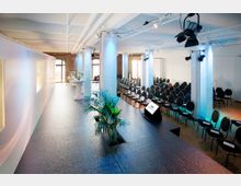 Moderner Veranstaltungsraum mit einer erhöhten Bühne, darauf Pflanzen und Präsentationsmonitore. Der Raum verfügt über Reihen schwarzer Stühle, runde Stehtische und weiß gestrichene Säulen; im Hintergrund große Fenster mit Tageslichteinfall.