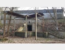 Teil eines verlassenen Geländes mit einer kleinen Betonstruktur und einem rostigen Überbau, dahinter ein großes Gebäude aus Wellblech. Die Umgebung ist von kahlen Bäumen und Laub bedeckt.