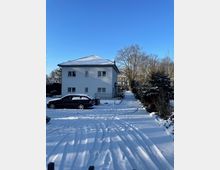 Verschneiter Innenhof mit breiter Zufahrt und Reifenspuren, links ein zweigeschossiges weißes Wohnhaus mit Walmdach und mehreren Fenstern. Rechts säumen Hecken und kahle Bäume den Weg; im Hintergrund weitere Wohngebäude unter blauem Himmel.