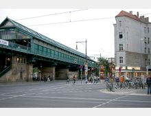 Straßenszene mit einer erhöhten, grünen U-Bahn-Station aus Metall und Glas; im Vordergrund eine Kreuzung mit Fußgängern, Fahrrädern und Verkehrszeichen. Rechts ein mehrstöckiges graues Gebäude mit Geschäften im Erdgeschoss.