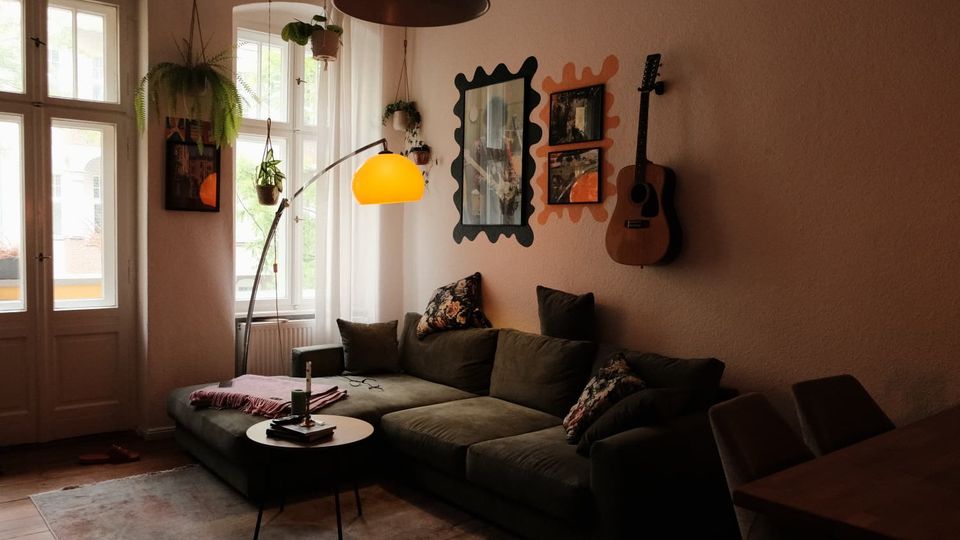 Gemütliches Wohnzimmer mit großer Eckcouch, rundem Couchtisch und Teppich vor hohen Altbaufenstern mit Balkontür. An der Wand hängen Spiegel und Bilder neben einer Gitarre; mehrere Hängepflanzen und eine Stehlampe mit gelbem Schirm prägen die Einrichtung.