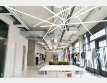 Moderner Showroom mit offener Gestaltung, weißen Wänden und einer Decke mit sichtbaren Belüftungsrohren und geometrischen Leuchtstäben. Im Raum stehen Kleiderstangen mit bunter Kleidung, große Fenster lassen Tageslicht herein, und zentral befinden sich ein Tisch mit Sitzgelegenheiten und eine grüne Couch.