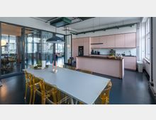 Moderne Gemeinschaftsküche mit pastellrosa Küchenzeile, schwarzer Arbeitsplatte und integrierten Geräten. Im Vordergrund steht ein großer Tisch mit transparenten Stühlen, darüber hängt eine schwarze Lampe; die Umgebung ist von Glaswänden und großen Fenstern geprägt.