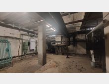 Kellerraum mit Rohrleitungen und technischen Installationen an Wänden und Decken. Der Raum hat eine rohe Betonoptik mit freiliegenden Rohren, einem Betonpfeiler, sowie einer alten Fensteröffnung im Hintergrund.