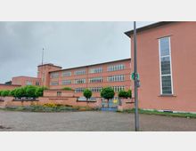 Backsteinfarbenes Schulgebäude mit flachem Dach, großen Fensterfronten und einem zentralen Turm mit Uhr. Vor dem Gebäude befinden sich ein Metalltor, eine gepflasterte Fläche und gepflegte Grünanlagen mit Blumenbeeten und kleinen Bäumen.