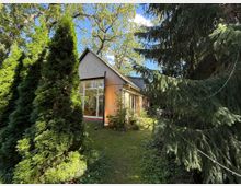 Ein modernes Haus mit einer Holzfassade und großen Fenstern, umgeben von hohen Nadelbäumen und einem gepflegten Garten. Im Hintergrund sind blauer Himmel und Laubbäume sichtbar.