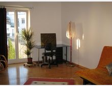 Helles Wohnzimmer mit Holzboden, einem Schreibtisch und Stuhl in der Ecke vor einer großen Fenstertür, die auf einen Balkon führt. Im Raum befinden sich zudem eine Topfpflanze, eine Stehleuchte, ein orangefarbenes Sofa und ein kleiner Teppich auf dem Boden.