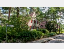 Ein Einfamilienhaus mit rotem Ziegeldach, roten Fensterläden und hellem Putz, umgeben von dichter Vegetation und hohen Bäumen. Im Vordergrund ein Straßenschild mit der Aufschrift "Himmelsteig" neben einem Gehweg.