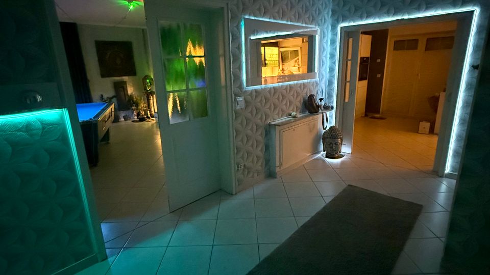 Innenansicht eines Flurs in einer Wohnung mit weiß gefliesten Böden, gemusterter Tapete und mehreren Türen zu angrenzenden Räumen. LED-Lichtleisten rahmen Tür- und Wandöffnungen, und an der Wand befinden sich ein Heizkörper sowie ein schmaler Teppichläufer auf dem Boden.