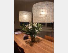 Ein Esszimmertisch aus Holz mit einem Blumenstrauß in einer Vase in der Mitte. Darüber hängen zwei große, quadratisch strukturierte Pendelleuchten, im Hintergrund ist ein Heizkörper vor einem Fenster sichtbar.