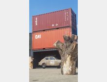 Gestapelte rote Frachtcontainer auf einem gepflasterten Platz, mit einem beigefarbenen Oldtimer-Auto (beschriftet mit der Nummer "62") davor und Baumstümpfen im Vordergrund. Der Himmel ist klar und blau.