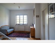 Schmaler, heller Wohnraum mit Holzfußboden, einem gemusterten Teppich und einem blau gestreiften Sofa an der linken Wand. Am Ende des Raums befindet sich ein mehrteiliges Fenster über einem Heizkörper; rechts stehen eine kleine Kommode und eine weiße Tür, an den Wänden hängen Bilder.