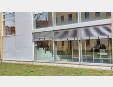 Moderne Gebäudefassade mit großen Glasfenstern, teilweise mit außenliegenden Jalousien ausgestattet. Im Vordergrund eine kleine Rasenfläche, im Hintergrund spiegeln sich Gebäude aus gelbem Backstein.
