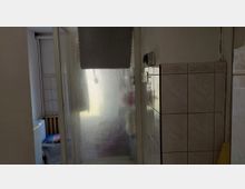 Kleines Badezimmer mit gefliester Dusche und milchiger Duschkabinentür, teilweise durch ein Wabenrollo abgedeckt. Links sind ein WC und ein Heizkörper zu sehen, rechts eine geflieste Trennwand mit Lichtschaltern.
