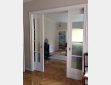 Ein Wohnraum mit einem hellen Holzboden in Fischgrätmuster, getrennt durch Schiebetüren mit Glasfenstern. Im Hintergrund ist ein Kamin, ein Regal mit modernen Dekorationen und ein weißer Teppich zu sehen.