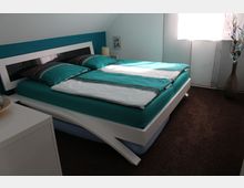 Modernes Schlafzimmer mit einem Doppelbett mit türkis-weißer Bettwäsche und einem weißen, geschwungenen Bettrahmen. Im Raum befinden sich ein Nachttisch, ein Fenster mit Tageslicht und eine dekorative Vase in einem minimalistischen Stil, auf braunem Teppichboden.