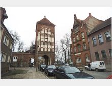 Straßenszene mit einem historischen Stadttor aus rotem Backstein in der Mitte, flankiert von Gebäuden im ähnlichen Stil. Im Vordergrund parken Autos auf einer gepflasterten Straße, während Bäume ohne Laub den Hintergrund säumen.