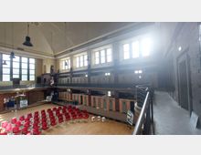 Innenraum eines historischen Gebäudes mit hohen Decken, großen Fenstern und einer Galerieebene. Der Raum enthält Reihen roter Stühle, eine Holzbodenfläche und Backsteinwände mit eingebauten Türen, die an Zellen erinnern.