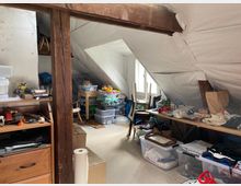 Ein Dachboden mit einer schrägen weißen Decke, Holzbalken und einem kleinen Fenster, durch das Tageslicht einfällt. Der Raum ist vollgestellt mit Regalen, Plastikkisten, Möbeln und verschiedenen Gegenständen wie Spielsachen, Kleidung und Haushaltswaren.