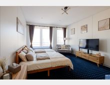 Helles Schlafzimmer mit einem Doppelbett aus Holz, einer grauen Polstercouch, einem großen Flachbildfernseher auf einem Holztisch und drei großen Fenstern mit weißen Vorhängen und dunklen Gardinen. Der Boden hat einen dunkelblauen Teppich, und an den Wänden hängen abstrakte Bilder.