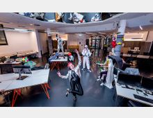 Großraumbüro mit modernen Schreibtischen und Computermonitoren, gestaltet mit auffälligen Kostümen und Requisiten von Personen, die kreativ posieren. Der Raum hat eine minimalistische Einrichtung mit weißen Wänden, dekorativen Säulen und einer Lichtdecke mit Spiegeln.