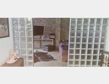 Wohnraum mit modernem und rustikalem Design; eine Wand aus Glasbausteinen trennt den Bereich mit nackter Ziegelwand, einem Fernseher, einem kleinen Tisch mit Kissen und einem Bett mit gemustertem Überwurf. Der Boden ist beige und mit einer dunklen Teppichauflage dekoriert.