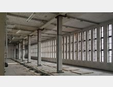 Großer, leerstehender Fabrikraum mit weiß gestrichenen Backsteinwänden, zahlreichen schmalen, hohen Fenstern und einer Decke mit freiliegenden Rohren und Leuchtstofflampen. Der Boden ist teilweise mit Holz und Trümmern bedeckt, und massive Säulen verlaufen in Reihen durch den Raum.