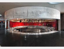 Runder, moderner Veranstaltungsraum mit Betonwänden und einer Bühne in der Mitte, umgeben von halbkreisförmigen Sitzreihen. Rote Vorhänge und Bühnenscheinwerfer dominieren den Innenraum, der durch die offene Architektur und dunkel glänzende Böden geprägt ist.