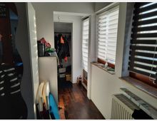 Schmaler, heller Flur mit dunklem Holzboden, rechts drei Fenster mit gestreiften Rollläden, links ein hoher Spiegel und ein kleiner Behälterschrank. Im Hintergrund ist ein Wanddurchgang zu sehen, der zu einer Garderobe mit Jacken und Schuhen führt.