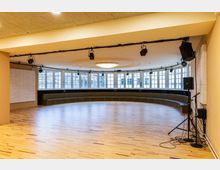 Großer, leerer Veranstaltungs- oder Probenraum mit hellem Holzboden, gebogener Fensterfront und umlaufender Sitzbank entlang der Wand. An der Decke sind Beleuchtungs- und Tontechnik montiert, rechts stehen Lautsprecher und Mikrofonstative.