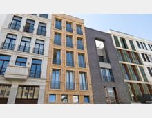 Mehrstöckige Wohngebäude in einem modernen Designstil, mit Fassaden aus hellem Putz, sandfarbenem Naturstein und dunklem Backstein. Die Fensterfronten sind symmetrisch angeordnet und teilweise mit dekorativen Balkongittern oder Fensterläden versehen.