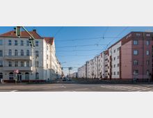 Straßenszene mit zwei gegenüberliegenden Wohngebäudereihen, links ein weißes Haus mit roten Dachziegeln, rechts eine Fassade in weiß und rosa mit Backsteinelementen. Oberhalb der Straße verlaufen zahlreiche Oberleitungen für Straßenbahnen, und es sind Ampeln sowie parkende und fahrende Autos sichtbar.