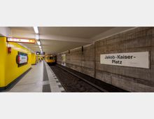 U-Bahn-Station Jakob-Kaiser-Platz mit gelb gekachelten Wänden und einem einfahrenden gelben Zug auf der rechten Gleisseite. Ein Stationsschild und Wegweiser hängen an den Wänden, während der Boden aus hellen Fliesen mit dunklen Streifen besteht.
